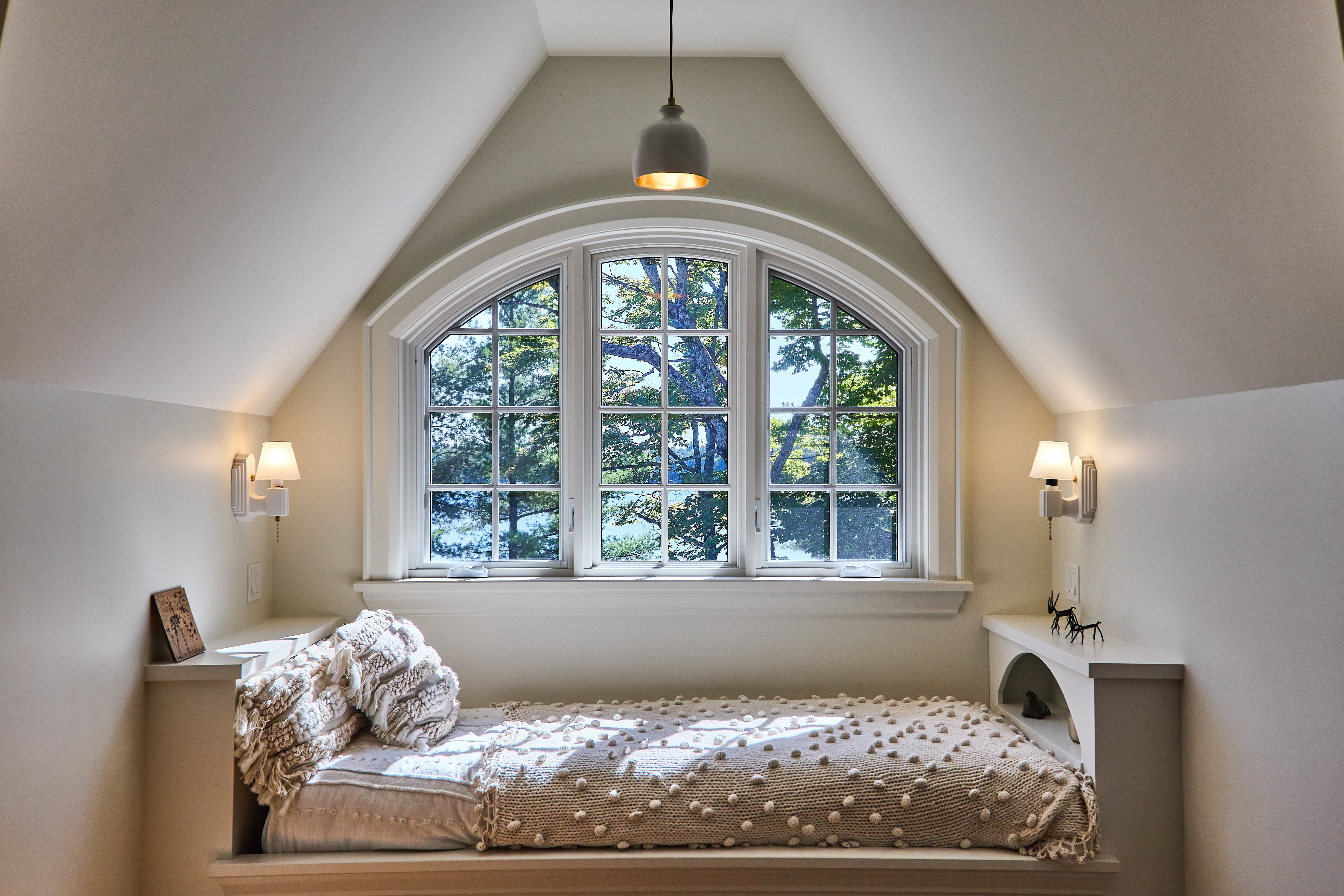 Cozy bedroom nook with a built-in bed under arched windows, framed by sloped ceilings and soft lighting.