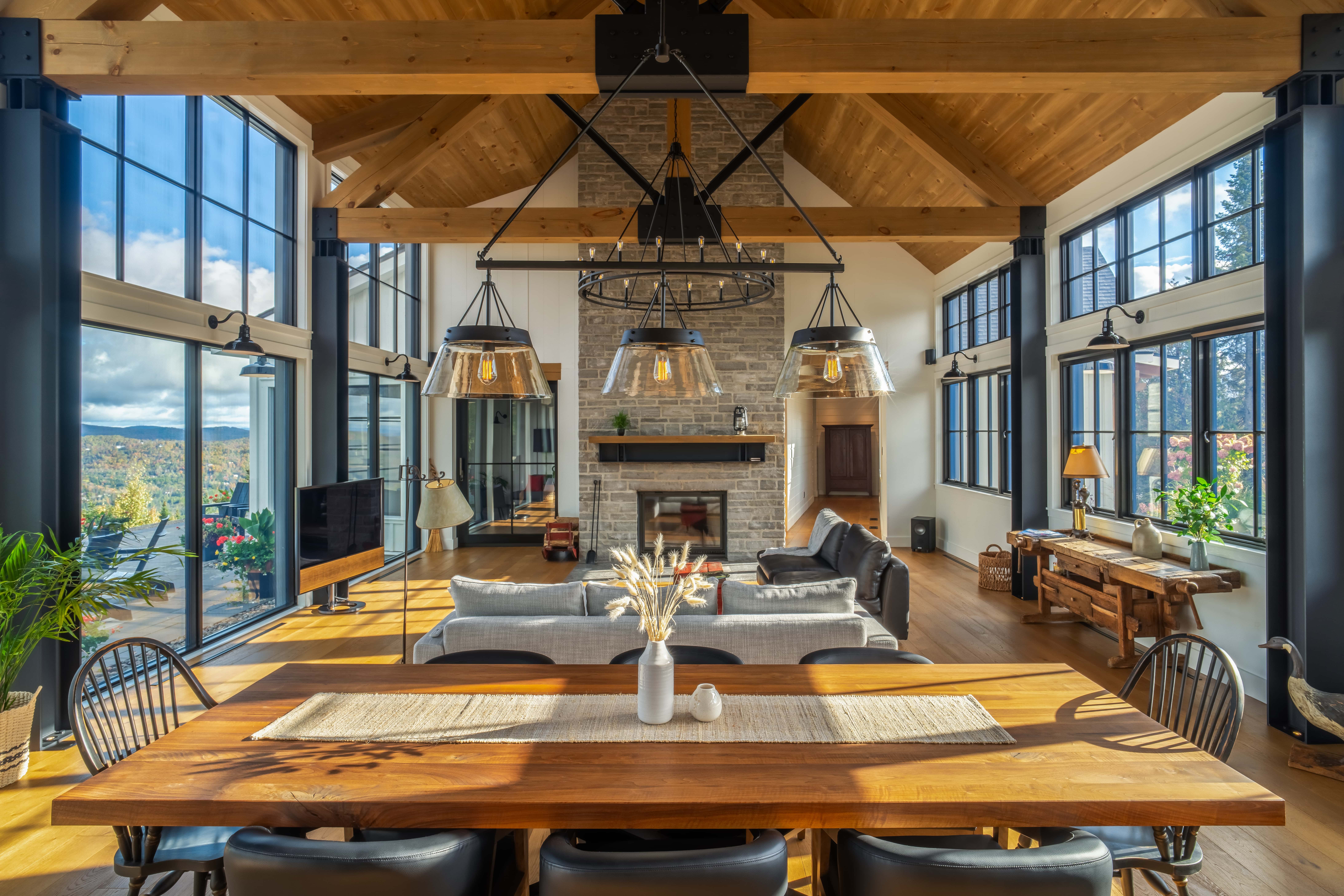 Rustic living room with stone fireplace, wooden beams, and floor-to-celing black-framed windows on either side.