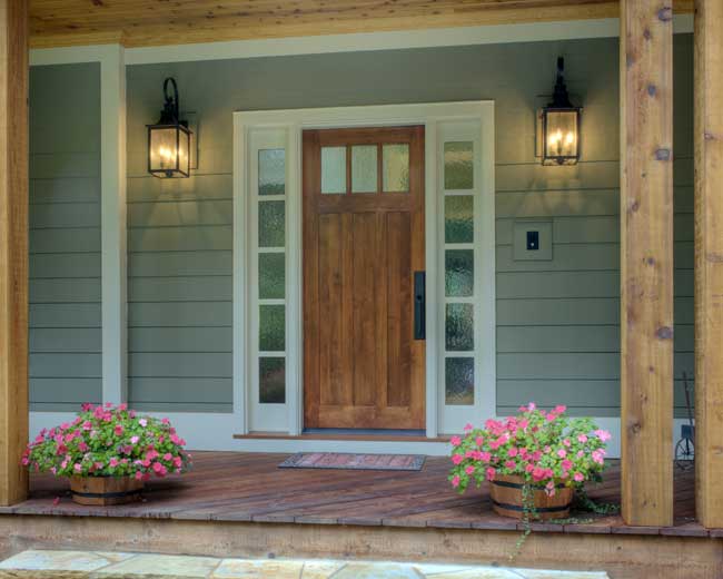 Wooden front door with sidelights, white trim, and lantern lights on a covered porch with flower planters.
